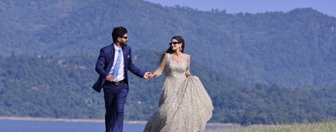 A couple in elegant wedding attire walking hand in hand by a picturesque riverbank in Una, India.